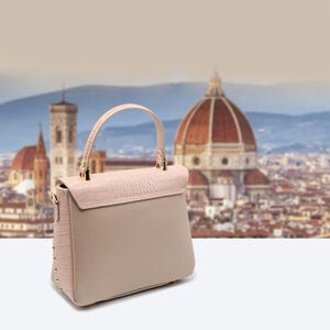 May include: A light pink and beige handbag with a top handle and a flap closure. The bag features a textured, crocodile-like pattern on the flap and side panels, with gold-colored studs and hardware. The background shows a blurred cityscape.