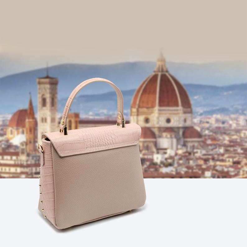 May include: A light pink and beige handbag with a top handle and a flap closure. The bag features a textured, crocodile-like pattern on the flap and side panels, with gold-colored studs and hardware. The background shows a blurred cityscape.