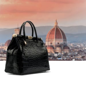 May include: A black, patent leather handbag with a crocodile-effect finish. The bag has two top handles and gold-tone metal clasps. The background shows a blurred cityscape with a large domed building.