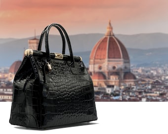 May include: A black, glossy handbag with a crocodile-skin texture. The bag has two handles and gold-colored hardware. The background features a blurred cityscape with a large domed building, suggesting a travel or fashion theme.