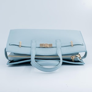 May include: A light blue leather handbag with gold-colored hardware. The bag features a top handle, a flap closure with a gold clasp, and a zippered compartment. The bag is made of textured leather and has a structured design.