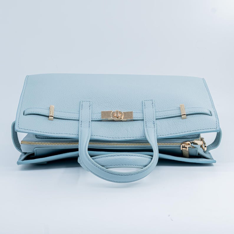 May include: A light blue leather handbag with gold-colored hardware. The bag features a top handle, a flap closure with a gold clasp, and a zippered compartment. The bag is made of textured leather and has a structured design.
