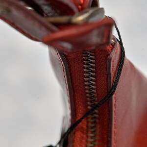 May include: Close-up of a red leather handbag with a zipper closure and a black tag attached.
