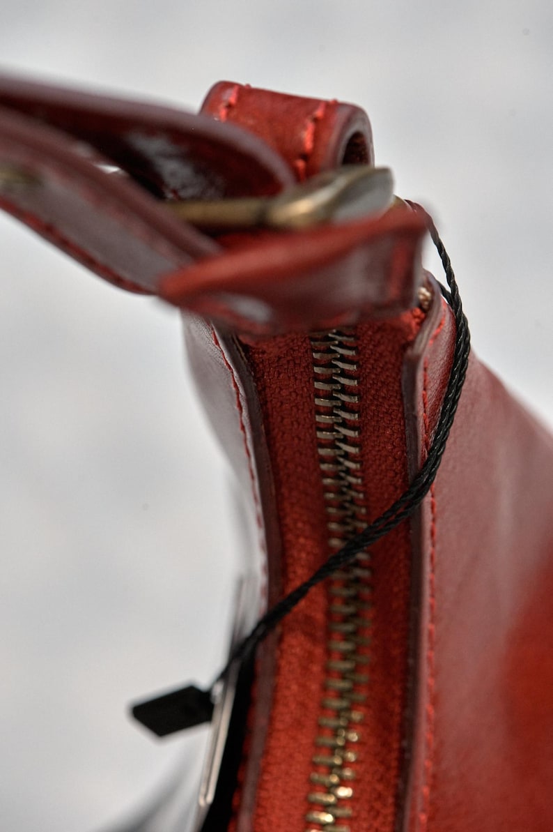 May include: Close-up of a red leather handbag with a zipper closure and a black tag attached.