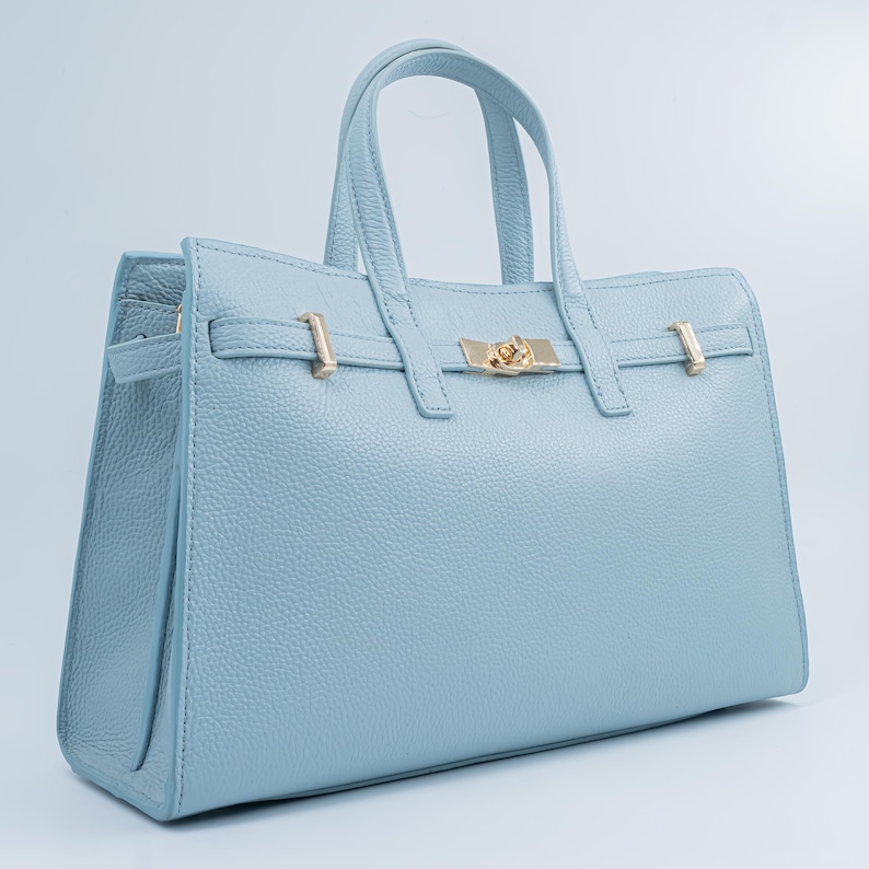 May include: A light blue leather handbag with a textured finish. The bag features two top handles, a gold-colored clasp, and gold-colored hardware. The handbag has a rectangular shape and a structured design.