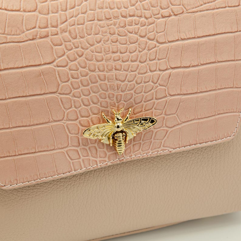 May include: A close-up of a light pink handbag with a textured, crocodile-like pattern. The flap features a gold-colored bee-shaped clasp. The lower portion of the bag is a smooth, lighter pink. The bag is a stylish accessory.