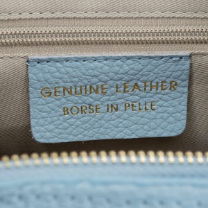 May include: Close-up of a light blue leather tag with the words "GENUINE LEATHER" and "BORSE IN PELLE" in gold lettering. The tag is stitched onto a light beige fabric, with a zipper visible at the top and bottom.