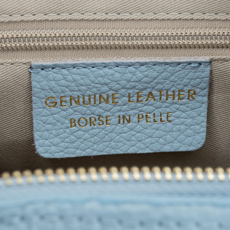 May include: Close-up of a light blue leather tag with the words "GENUINE LEATHER" and "BORSE IN PELLE" in gold lettering. The tag is stitched onto a light beige fabric, with a zipper visible at the top and bottom.