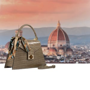 Peut inclure: Un sac à main taupe avec une texture crocodile, une poignée supérieure et un foulard décoratif. Le sac a un fermoir doré et une bandoulière assortie. L'arrière-plan présente un paysage urbain flou avec un grand bâtiment en forme de dôme.