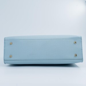 May include: A light blue handbag with a textured surface. The bottom of the bag features four gold-colored metal feet. The bag has a rectangular shape and a stitched seam along the top.