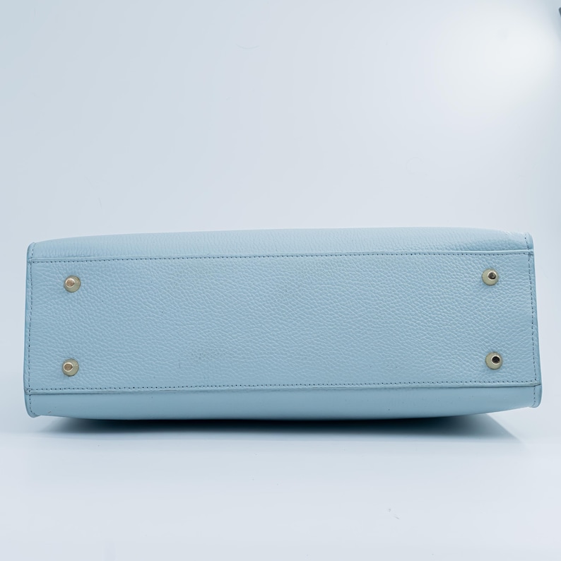 May include: A light blue handbag with a textured surface. The bottom of the bag features four gold-colored metal feet. The bag has a rectangular shape and a stitched seam along the top.