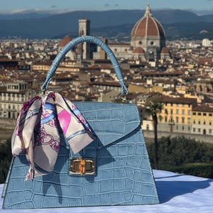 Peut inclure: Un sac à main bleu clair en cuir gaufré crocodile avec une poignée supérieure et un foulard assorti à motif floral rose et bleu. Le sac est devant un paysage urbain avec un grand bâtiment en forme de dôme en arrière-plan.