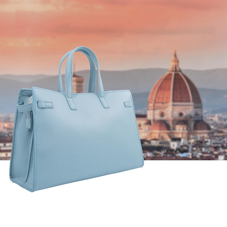 May include: A light blue leather handbag with two top handles and a structured design. The bag features a zipper closure and a rectangular shape. The background shows a blurred cityscape with a large dome.