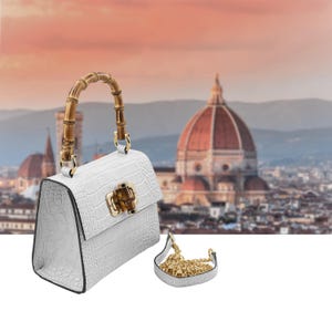 May include: White handbag with a bamboo handle and gold-tone hardware. The bag has a crocodile-like texture and a matching small chain strap. The background features a blurred cityscape with a large domed building.