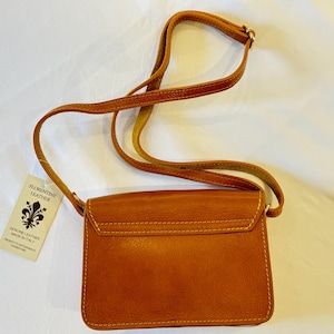 May include: A brown leather crossbody bag with a flap closure and a gold metal accent. The bag has a small tag attached to it that reads "Florentine Leather, Genuine Leather Made in Italy".