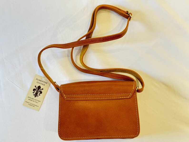 May include: A brown leather crossbody bag with a flap closure and a gold metal accent. The bag has a small tag attached to it that reads "Florentine Leather, Genuine Leather Made in Italy".