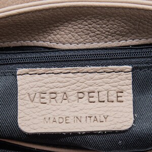 May include: Close-up of a beige leather bag with a black zipper. A beige leather tag is stitched to the bag and reads "VERA PELLE MADE IN ITALY" in embossed lettering. The leather has a textured appearance.