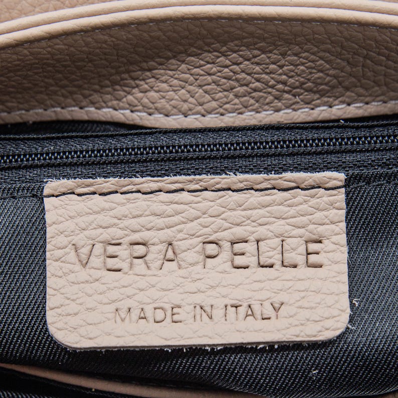May include: Close-up of a beige leather bag with a black zipper. A beige leather tag is stitched to the bag and reads "VERA PELLE MADE IN ITALY" in embossed lettering. The leather has a textured appearance.