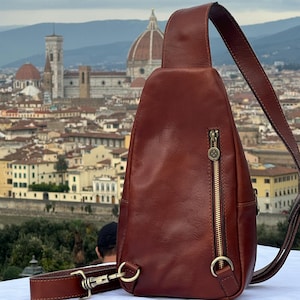 May include: A brown leather crossbody bag with a gold zipper and a silver buckle. The bag is worn over the shoulder and has a strap that can be adjusted. The bag is in front of a cityscape.