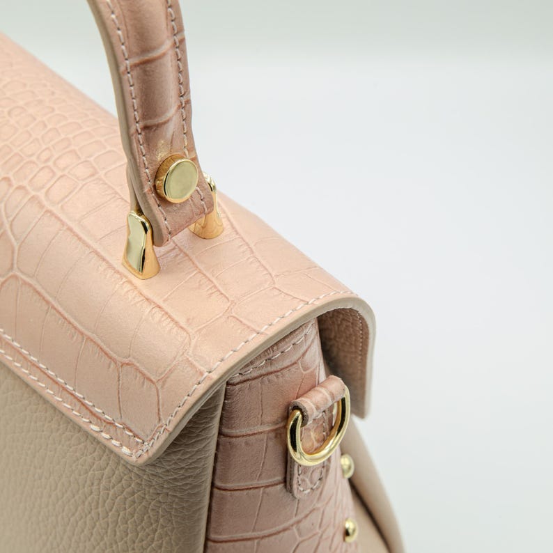 May include: A close-up of a light pink handbag with a textured, crocodile-like pattern. The bag features a top handle and gold-colored hardware. The bag's design includes a flap closure and a shoulder strap attachment.