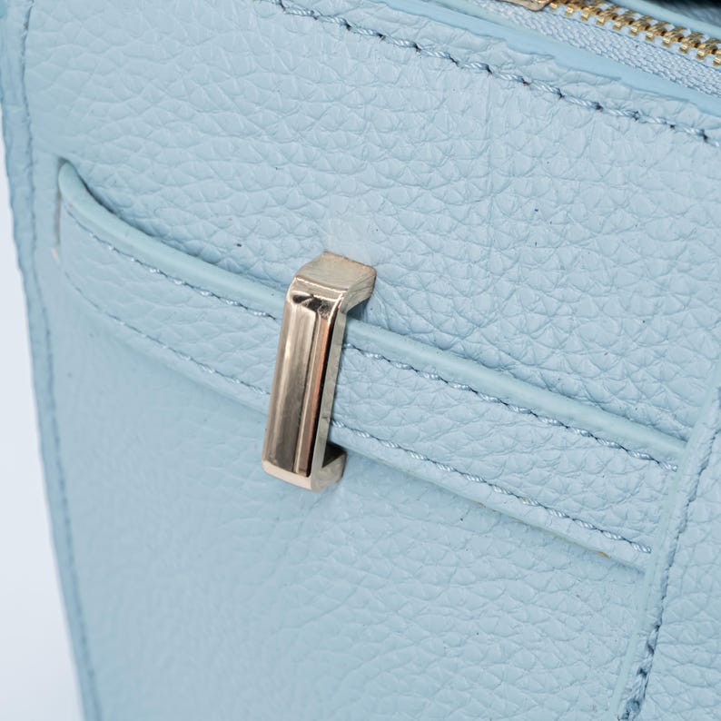 May include: Close-up of a light blue handbag with a textured leather finish. The bag features a gold-colored metal clasp and zipper. The stitching is visible along the edges of the bag and pocket.