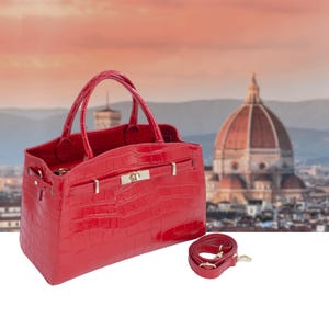 Peut inclure: Un sac à main en cuir rouge avec une texture peau de crocodile. Le sac a deux poignées et une bandoulière amovible. Le sac a un fermoir et des accessoires argentés. L'arrière-plan présente un paysage urbain flou.