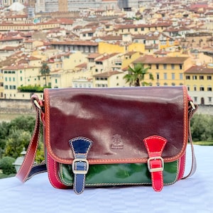 Peut inclure: Un sac à bandoulière en cuir marron avec des accents verts, rouges et bleus. Le sac a deux boucles et une étiquette de marque qui indique "Firenze Made in Italy".