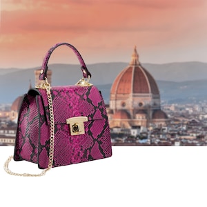 Puede incluir: Un bolso magenta con un estampado de piel de serpiente y una correa de cadena dorada. El bolso tiene un asa superior y un cierre de broche dorado. El fondo presenta un paisaje urbano borroso con un gran edificio abovedado.
