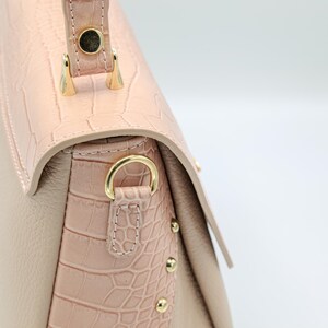 May include: Close-up of a beige and pink leather handbag. The bag features a textured, crocodile-like pattern on the flap and side panels. Gold-colored hardware, including a handle attachment and decorative studs, adds detail to the design.