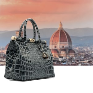 May include: A grey, crocodile-effect handbag with gold-coloured hardware, including a clasp and a decorative charm. The bag has two handles and a structured shape. The background features a blurred cityscape with a large domed building.