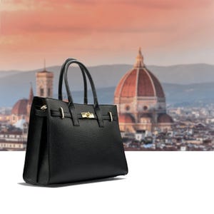 May include: A black leather handbag with a structured design, featuring a top handle and a gold-tone clasp. The bag has a rectangular shape and a textured surface. The background shows a cityscape with a large dome.