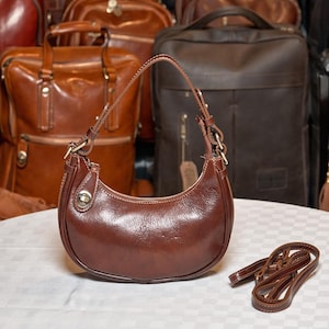 May include: A brown leather handbag with a curved shape and a long strap. The bag has a silver buckle and a silver button on the front.