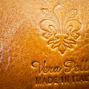 May include: Close-up of a brown leather tag with a fleur-de-lis design and the text "Vera Pelle Made in Italy".