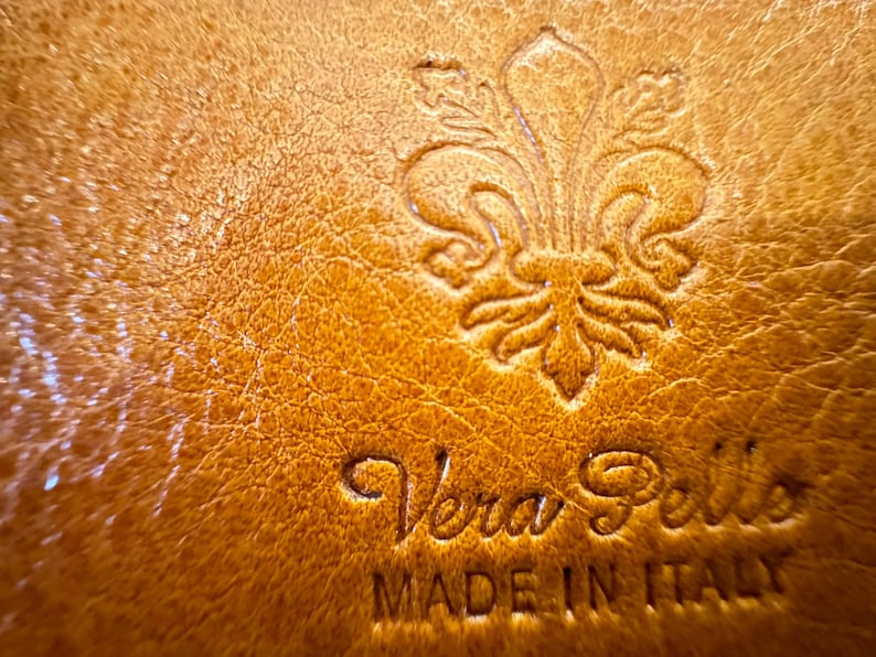May include: Close-up of a brown leather tag with a fleur-de-lis design and the text "Vera Pelle Made in Italy".