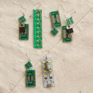 Puede incluir: Pendientes hechos a mano con componentes electrónicos reciclados. Los pendientes muestran placas de circuito verde, pequeños componentes cuadrados y un elemento de bombilla. Cada pendiente tiene un gancho plateado.