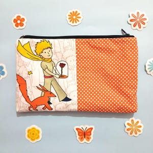 May include: A white and orange zippered pouch with a print of The Little Prince holding a rose in a glass dome and a fox walking beside him. The other side of the pouch is orange with white polka dots.