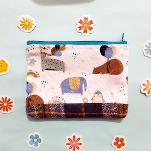 May include: A pink zippered pouch with a brown plaid trim. The pouch features a whimsical design with animals and balloons. The animals include an elephant, a bear, and a penguin.