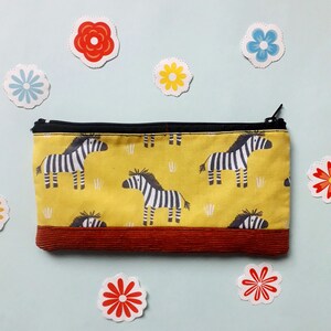 May include: A yellow pencil case with a brown corduroy bottom. The case is decorated with a repeating pattern of black and white zebras.