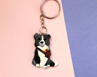 May include: A black and white border collie dog keychain with a brown satchel. The dog is sitting with its tongue sticking out and has a friendly expression. The keychain is attached to a silver key ring.