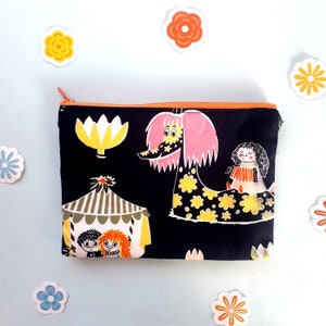 May include: Black zippered pouch with a colorful print featuring a pink and white creature with black eyes, a yellow flower, a green and white tent, and two figures with brown hair. The print is on a black background.