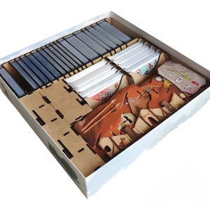 May include: A board game organizer with various compartments. The box contains stacks of cards, wooden dividers, and game pieces. The organizer is made of light brown wood and is designed to hold the game components neatly. The box is white with a landscape design.