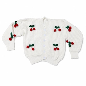 May include: A white long-sleeve knit cardigan featuring a V-neckline and a front button closure. The garment is decorated with multiple three-dimensional red cherry appliques, each paired with small green fabric leaves scattered across the front.