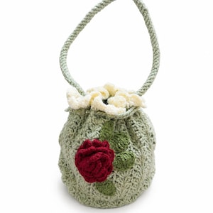 May include: A sage green crochet drawstring bag featuring a textured shell stitch pattern, a single handle, and a ruffled cream-colored top edge. The front is decorated with a dark red crocheted rose and two small green crocheted leaves.