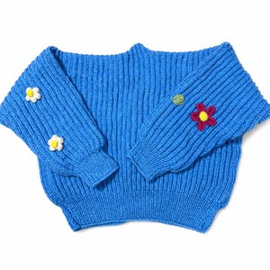 May include: A bright blue ribbed knit sweater featuring long sleeves with small, hand-stitched floral appliqu&eacute;s. The left sleeve displays two white flowers with yellow centers, while the right sleeve features one green leaf and one dark pink flower.