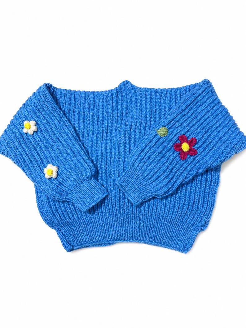 May include: A bright blue ribbed knit sweater featuring long sleeves with small, hand-stitched floral appliqu&eacute;s. The left sleeve displays two white flowers with yellow centers, while the right sleeve features one green leaf and one dark pink flower.