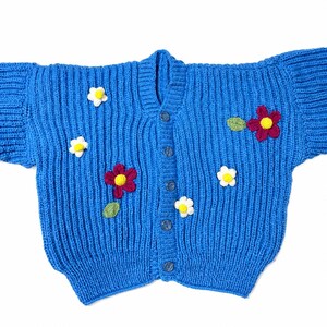May include: A bright blue knitted cardigan featuring a vertical ribbed texture and a front button closure. The garment is decorated with several small, three-dimensional crocheted flowers in white and deep red, each with a yellow center.