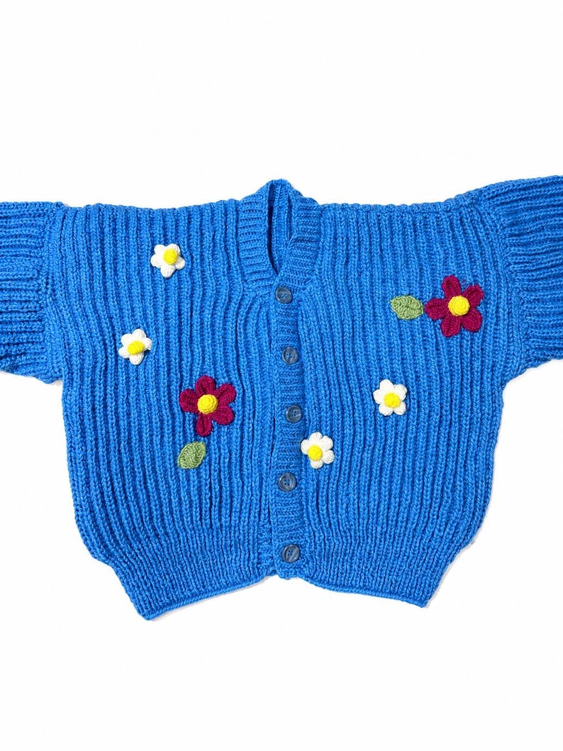 May include: A bright blue knitted cardigan featuring a vertical ribbed texture and a front button closure. The garment is decorated with several small, three-dimensional crocheted flowers in white and deep red, each with a yellow center.