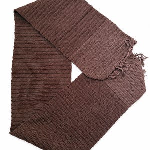 May include: A long, rectangular scarf in a solid dark brown color. The scarf features a consistent ribbed knit texture throughout its length and is finished with short, frayed fringe detailing along the edges of one end of the fabric.