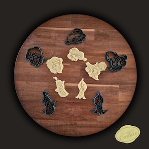 Pode incluir: Uma coleção de cortadores de biscoitos com rostos de personagens de desenhos animados, incluindo uma princesa, um gênio e um príncipe. Os cortadores são pretos e bege, dispostos em uma tábua redonda de madeira. Um adesivo "Design Cookie" está no canto inferior direito.
