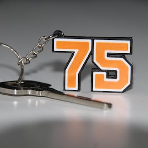 May include: A keyring with a silver key and a charm displaying the number 75 in orange with a white outline and black border. The charm is connected to a silver chain and ring.
